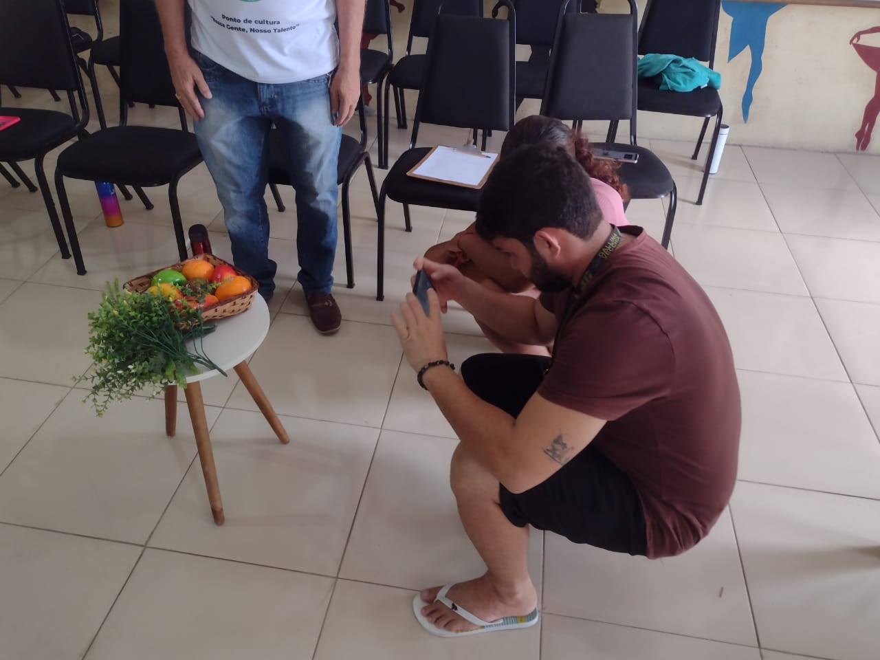 Homem agachado fotografando uma cesta de frutas e uma planta sobre mesa branca, com camisa do Ponto de Cultura ao fundo.