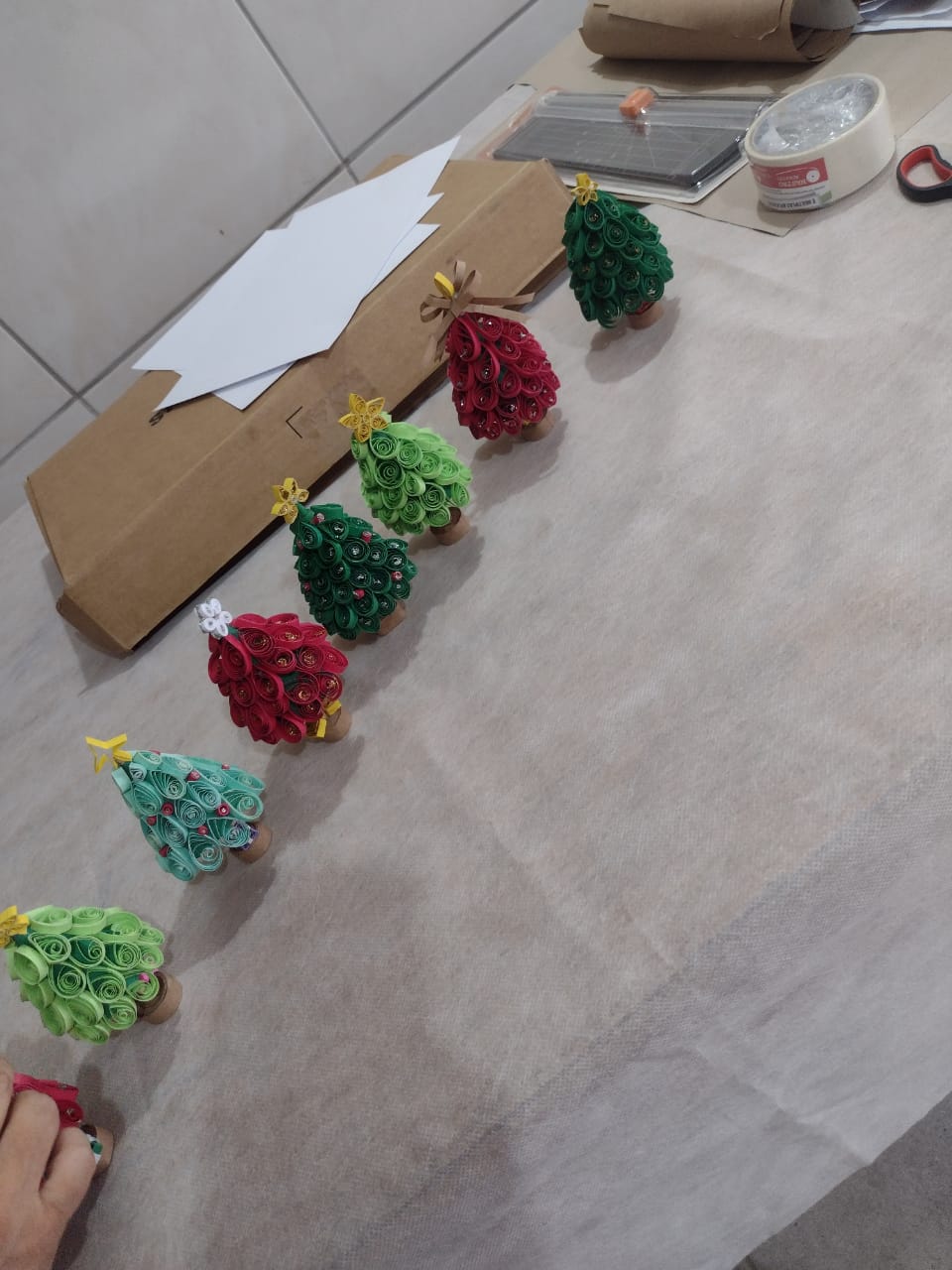 Árvores de Natal pequenas feitas em quilling, de papel enrolado em verde, vermelho e turquesa, sobre a mesa da oficina.