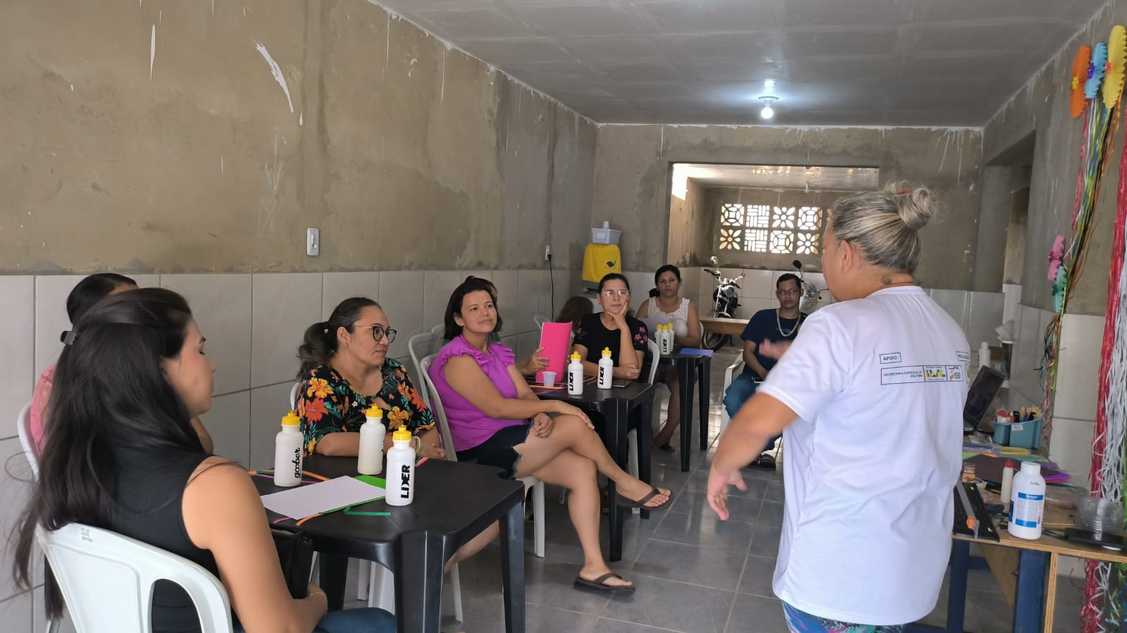 Grupo em treinamento da oficina de quilling, sentado em cadeiras ouvindo a instrutora na sala de artesanato.